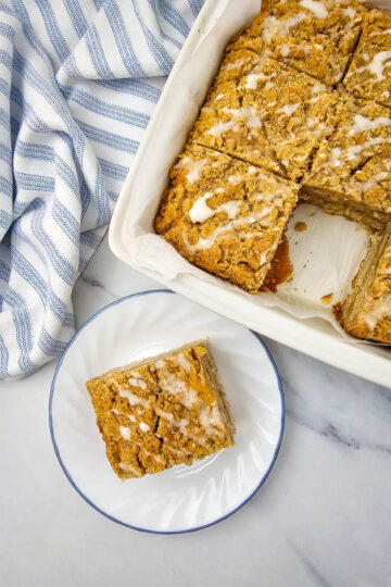 apple crumb cake in a pan with one slice cut out and placed on a white plate.