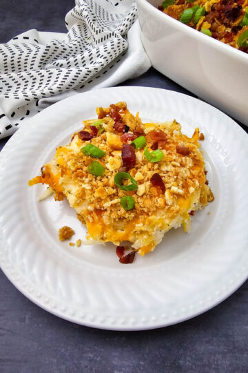 Serving of Million Dollar Potato Casserole on a white plate with buttery cracker topping, bacon pieces, and green onions.