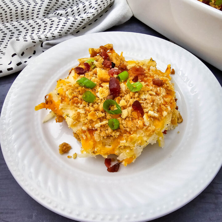 Serving of Million Dollar Potato Casserole on a white plate with buttery cracker topping, bacon pieces, and green onions.
