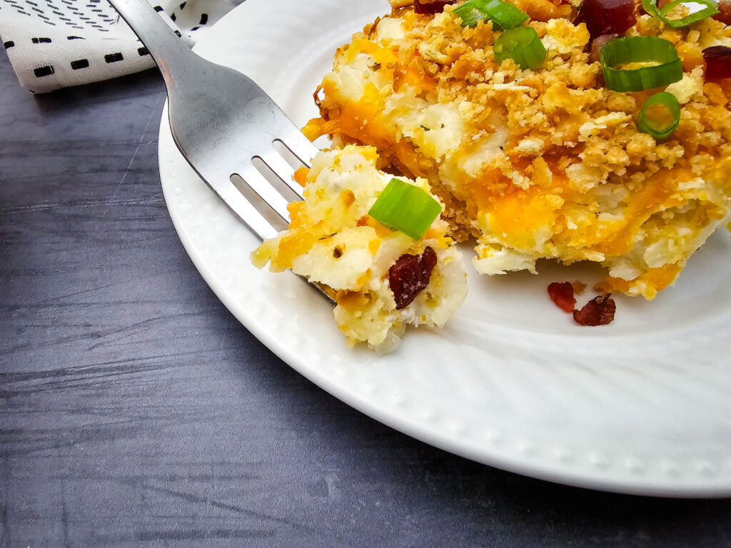 Close-up of creamy potato casserole with cheddar cheese, cracker topping, bacon, and green onions on a fork.