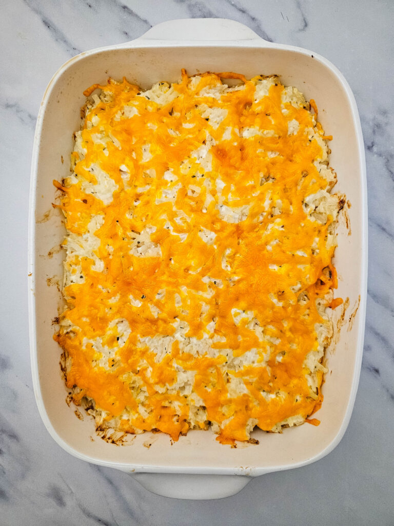 Baked hashbrown casserole topped with melted cheddar cheese in a white casserole dish.