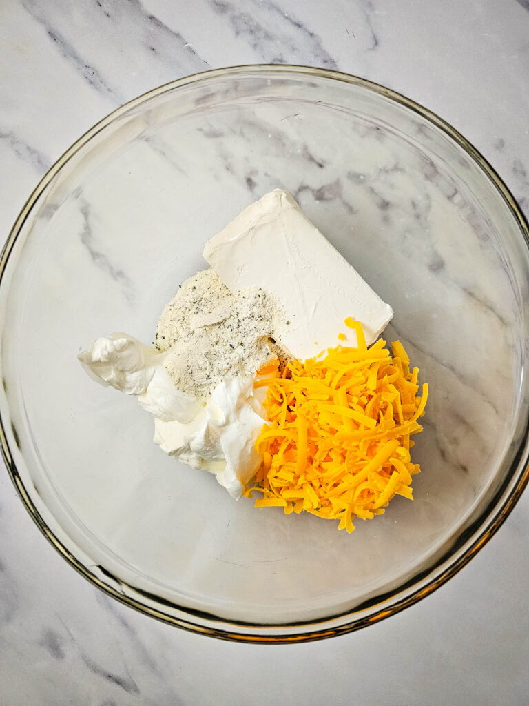 Cream cheese, sour cream, ranch seasoning, and shredded cheddar cheese in a glass mixing bowl.