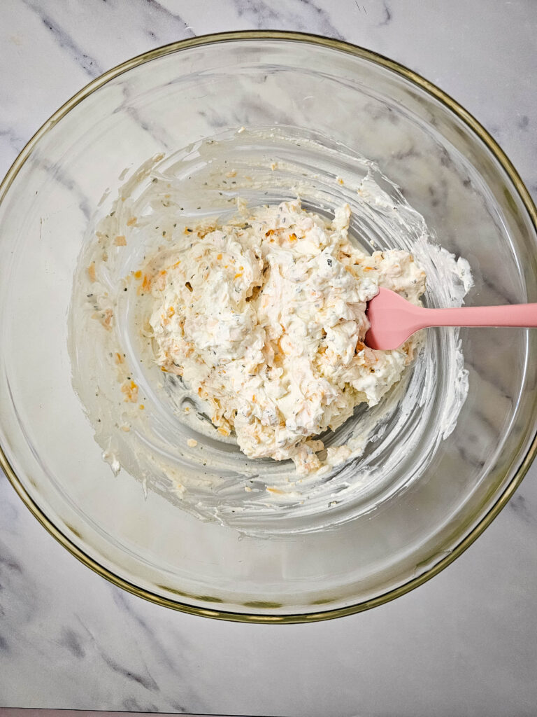 Cream cheese, sour cream, ranch seasoning, and cheddar cheese mixture stirred together in a glass bowl.