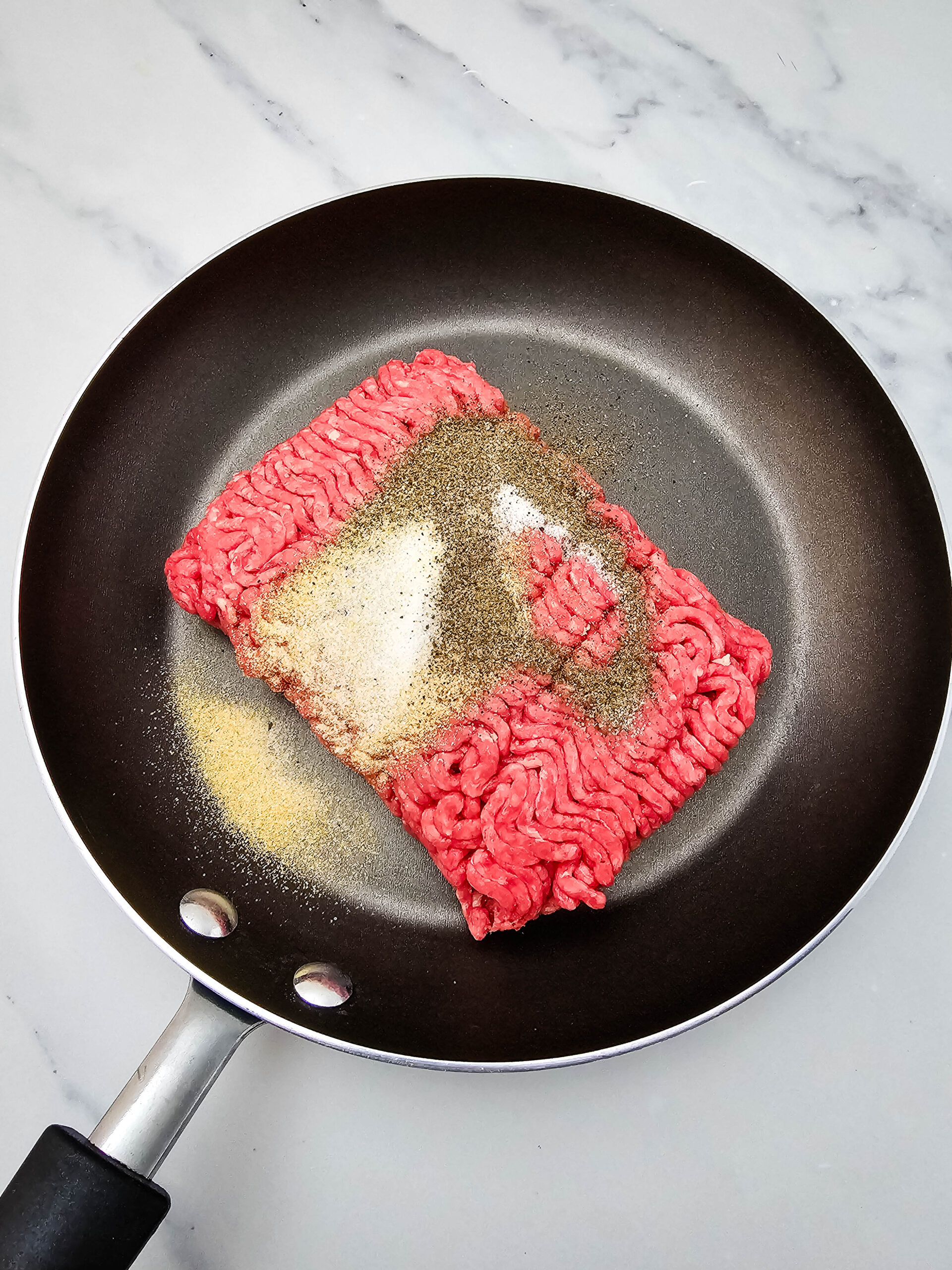 Ground beef in a skillet topped with garlic powder, salt, and pepper for Hobo Casserole.