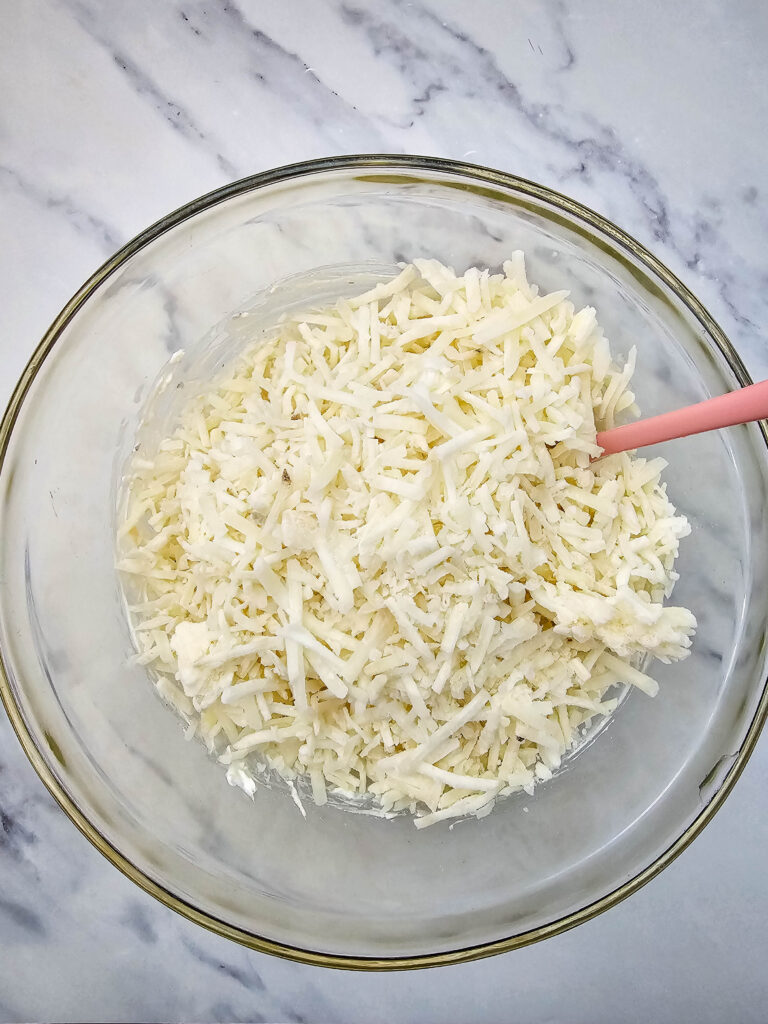 Frozen shredded hashbrowns in a glass mixing bowl with a pink spatula.