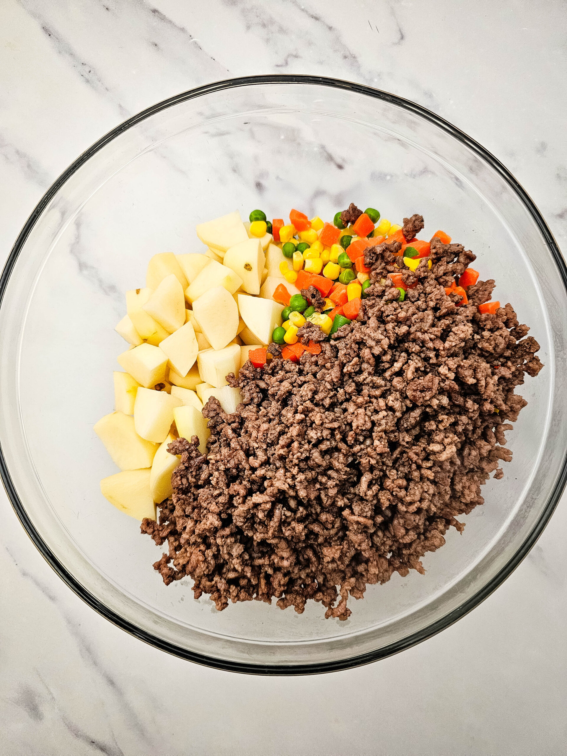 Cooked ground beef added to a glass bowl with diced potatoes and mixed vegetables for Hobo Casserole.