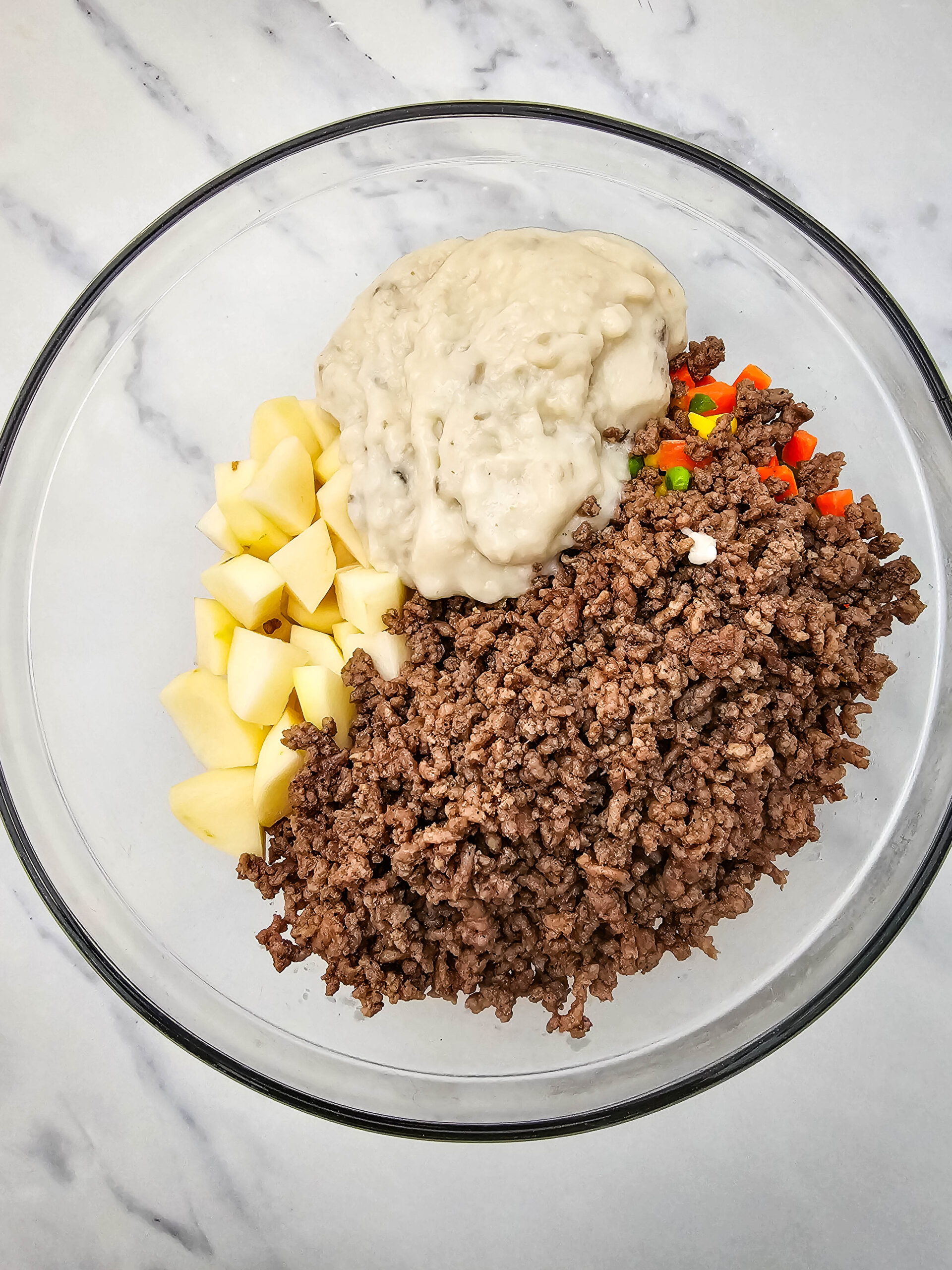 Cream of mushroom soup added to cooked ground beef, potatoes, and mixed vegetables in a glass bowl.