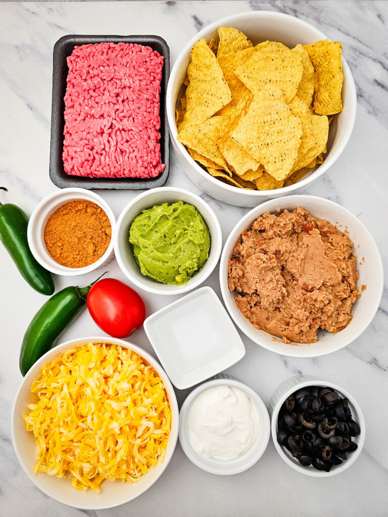 Ground beef nachos ingredients arranged on a marble counter, including tortilla chips, ground beef, cheese, beans, jalapeños, tomato, sour cream, olives, guacamole, and taco seasoning.