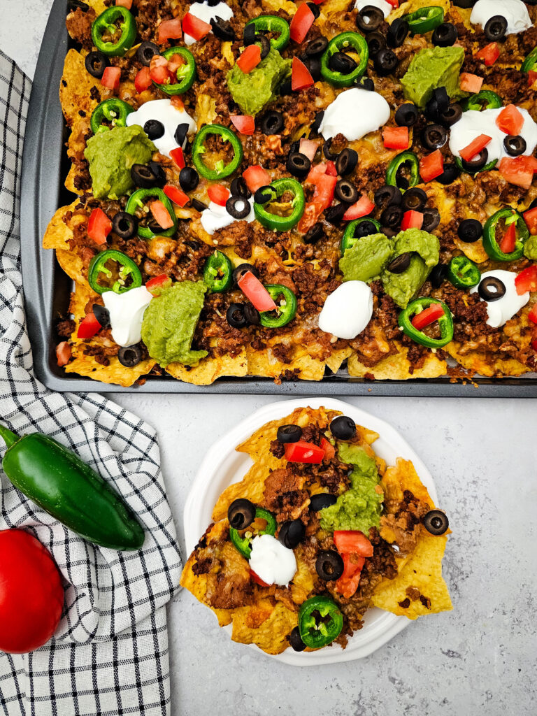 Loaded sheet pan ground beef nachos topped with beef, cheese, jalapeños, olives, tomatoes, sour cream, and guacamole, with a serving on a small plate.