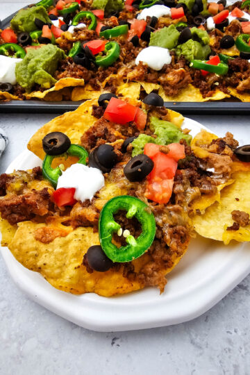 Sheet pan ground beef nachos served on a white plate with jalapeños, tomatoes, olives, sour cream, and guacamole.
