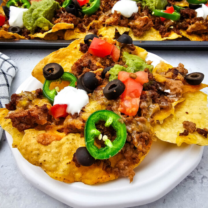 Sheet pan ground beef nachos served on a white plate with jalapeños, tomatoes, olives, sour cream, and guacamole.