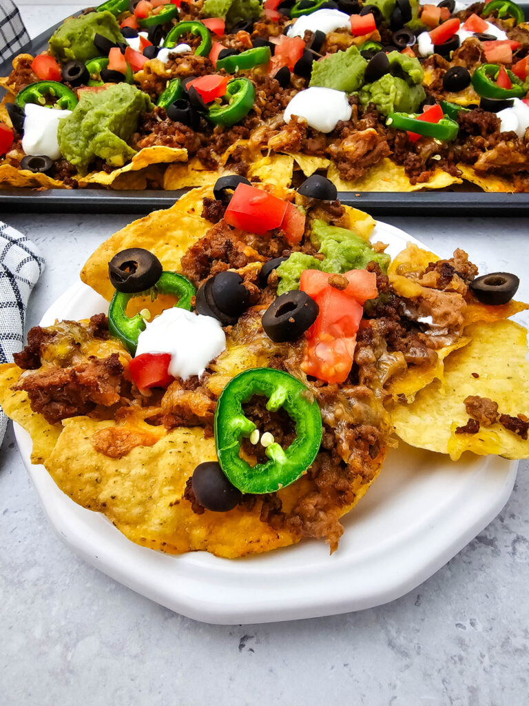 Sheet pan ground beef nachos served on a white plate with jalapeños, tomatoes, olives, sour cream, and guacamole.
